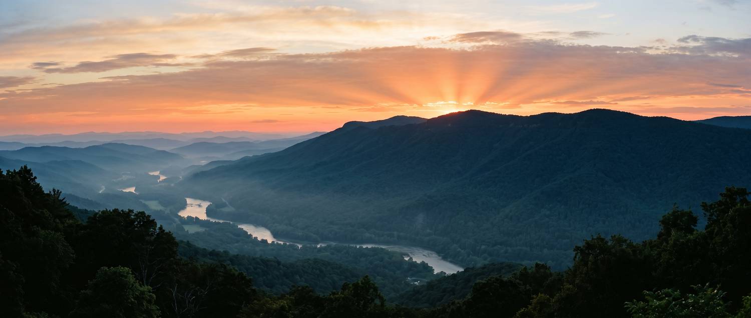 The sun setting behind a forested mountain ridge above a hazy river valley